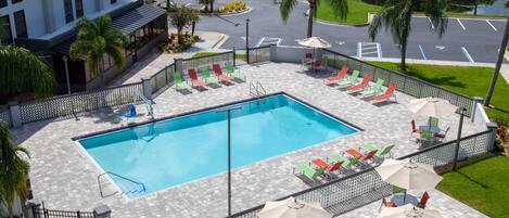 A swimming pool with lounge chairs and umbrellas, surrounded by a fence and palm trees.