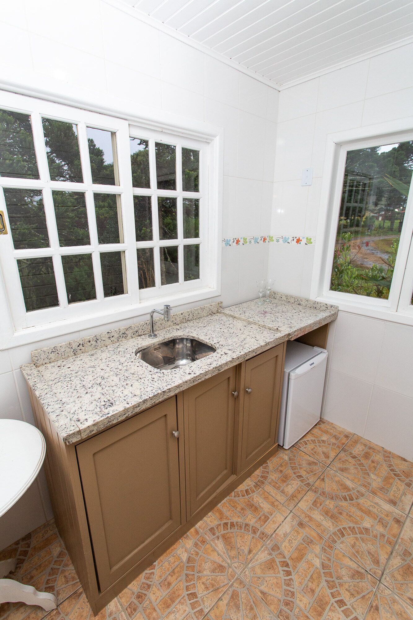 A kitchen with a granite countertop, a stainless steel sink, and a refrigerator. There are two windows with white frames, one of which has a view of greenery outside.
