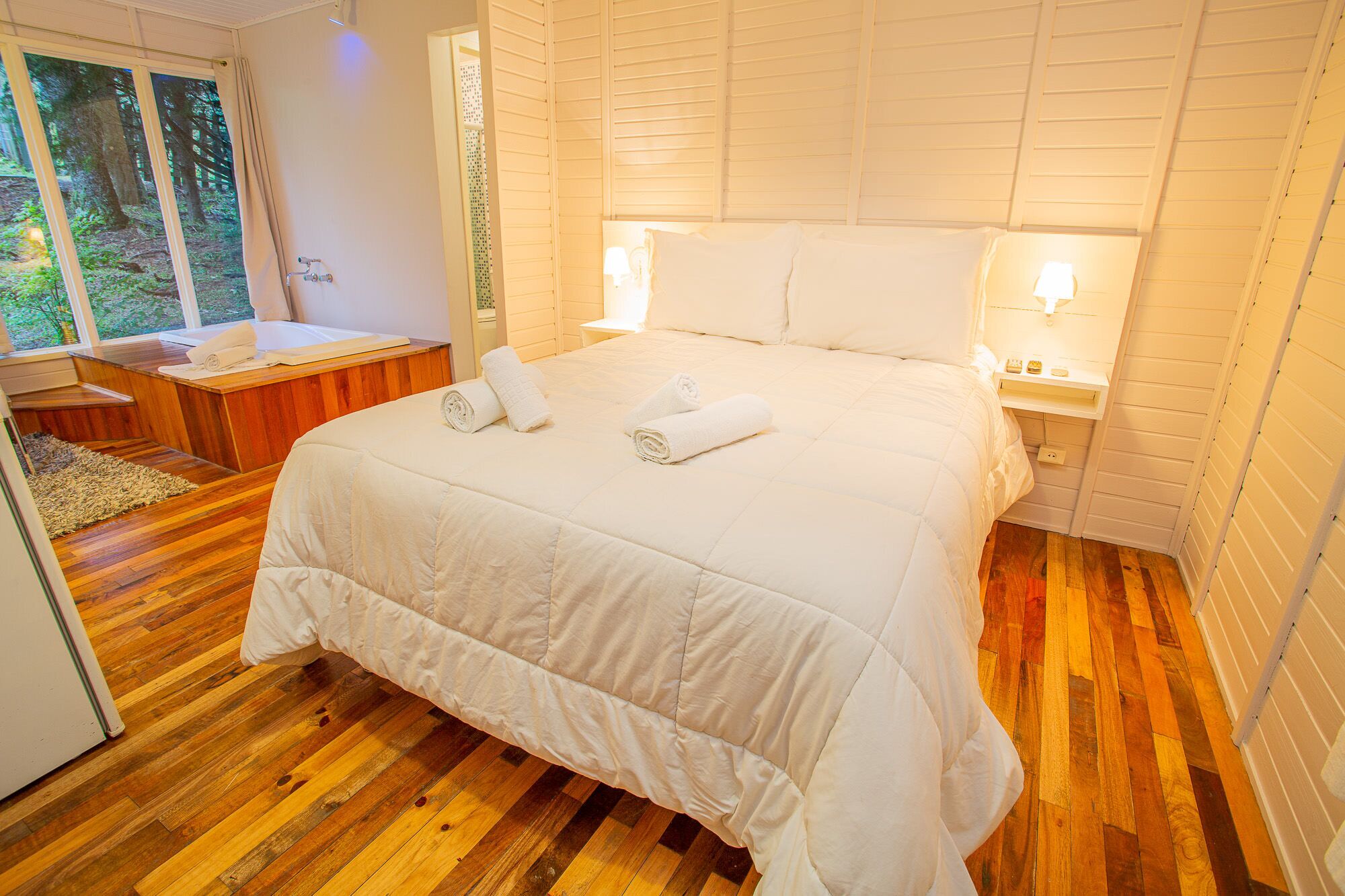 Een slaapkamer met een groot bed, twee bedlampjes en uitzicht op een bos door een raam.