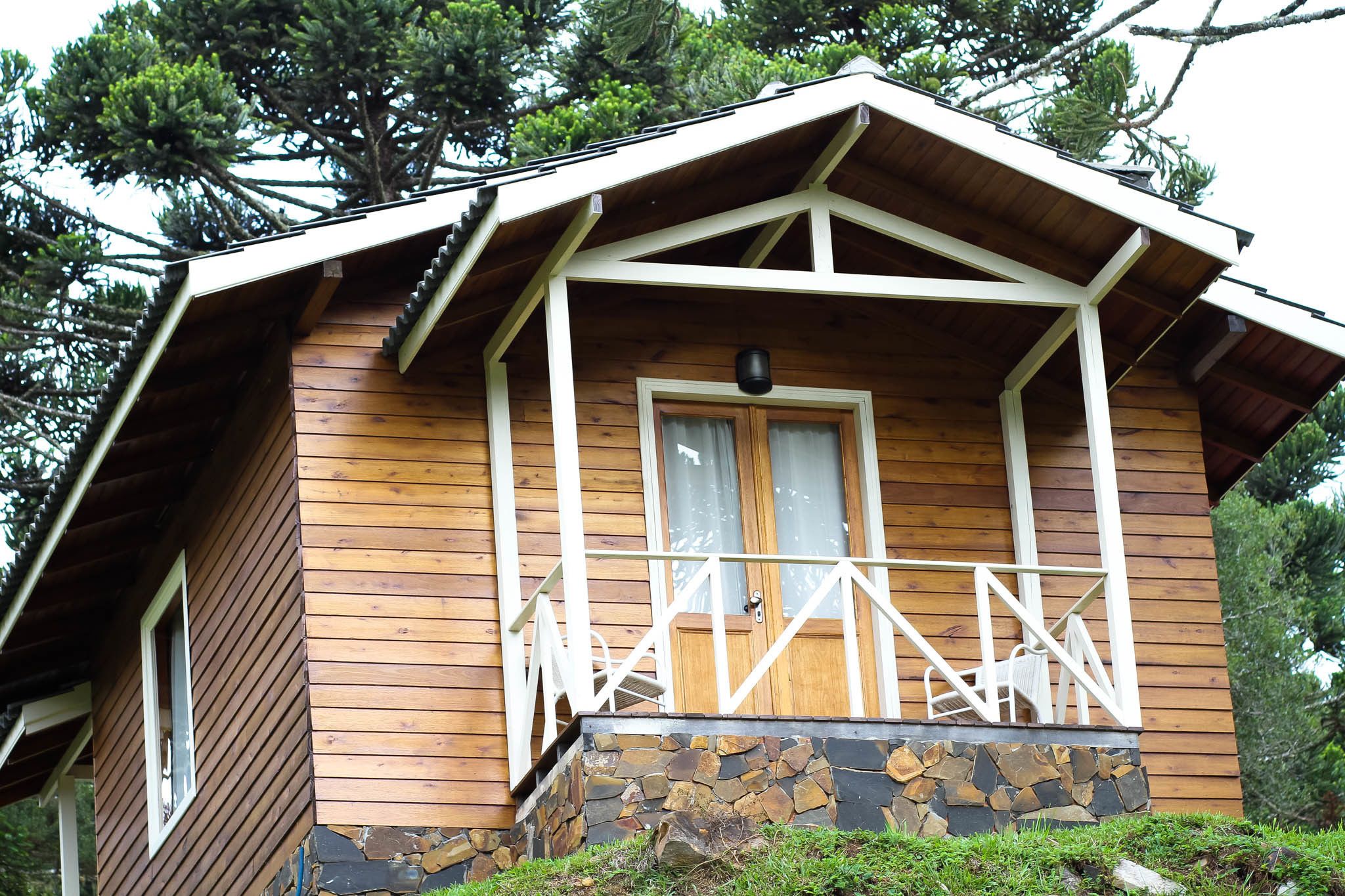 En stuga byggd av trä med en grund av sten, en veranda med vita räcken och ett fönster med gardiner.