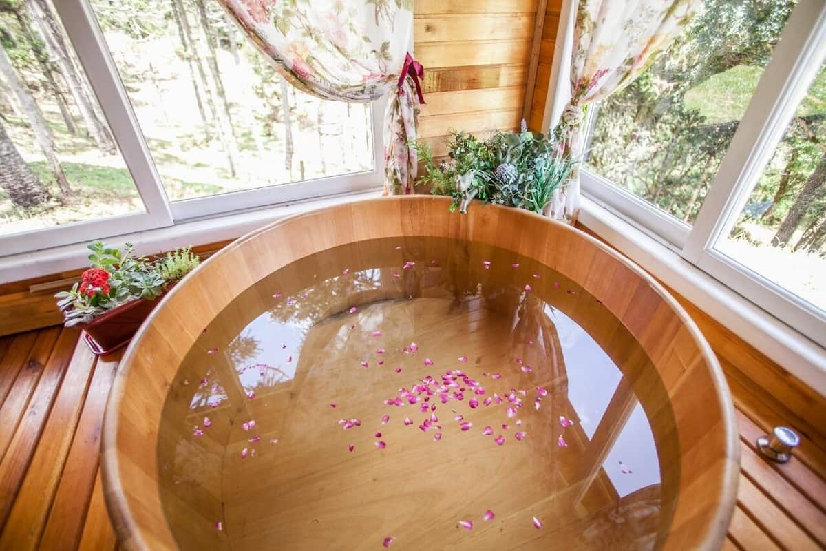 Une baignoire en bois remplie d’eau et de pétales de rose, de grandes fenêtres avec des rideaux et une vue sur les arbres.