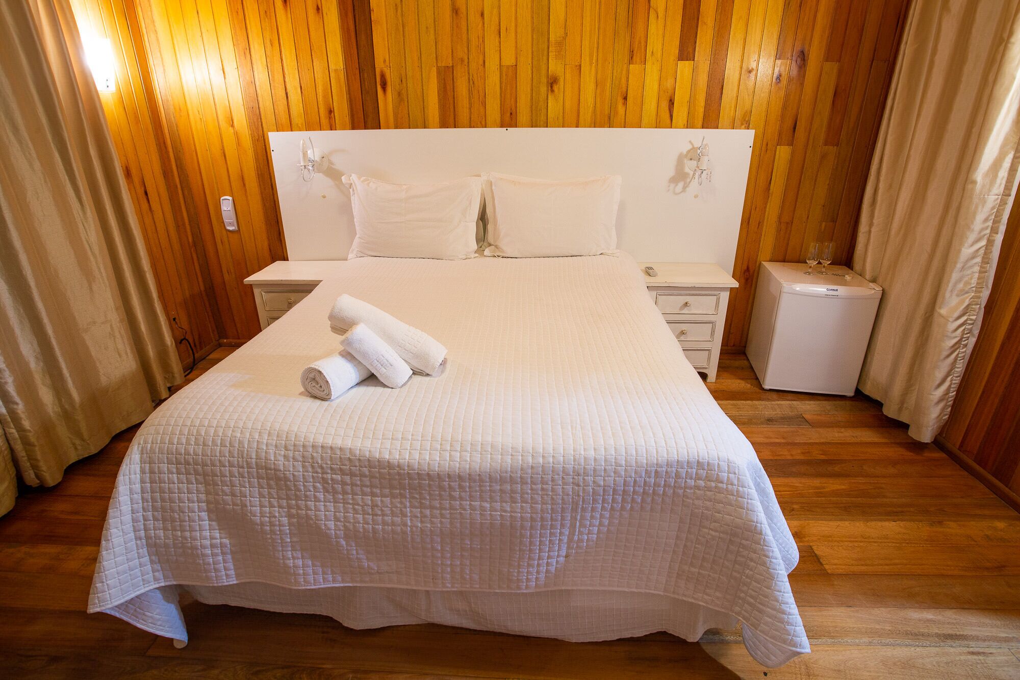 Un lit bien fait, avec du linge de lit blanc et des serviettes pliées, une table de chevet avec une lampe, une petite table de chevet avec un téléphone et un petit réfrigérateur.