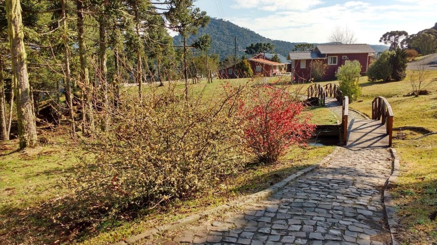 A cobblestone pathway leading to a rustic wooden bridge over a small stream, surrounded by lush greenery and a backdrop of mountains.