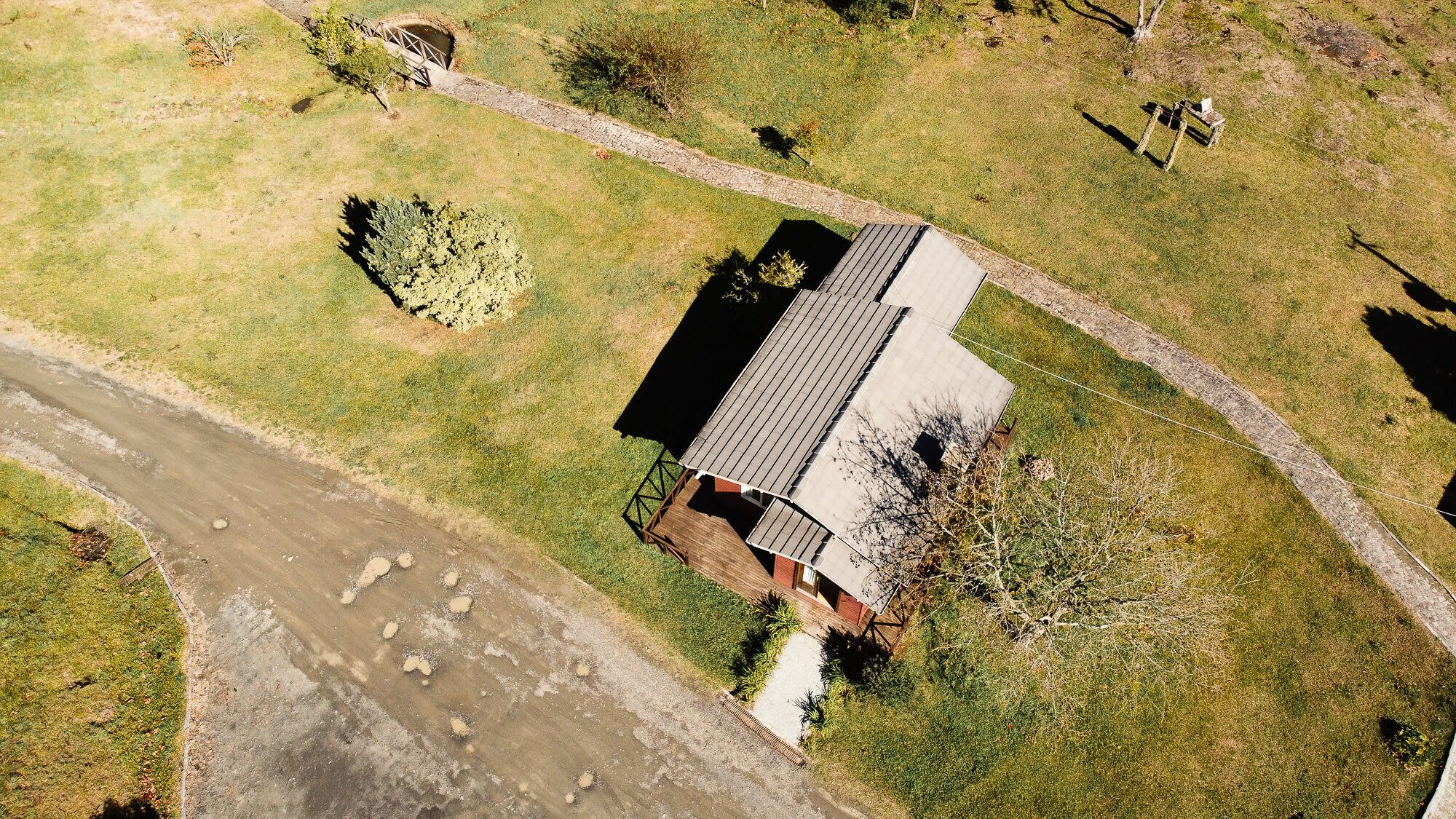 An aerial view of a single-story house with a sloped roof, surrounded by a grassy area with a few trees and a stone pathway.