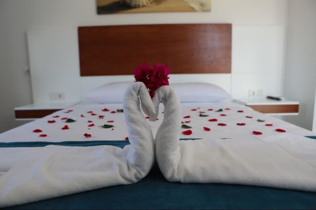 A neatly made bed with a white blanket and red rose petals scattered across it.