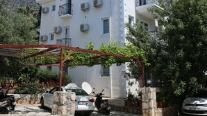 Un edificio blanco de varios pisos con balcones, coches aparcados y un muro de piedra al frente.