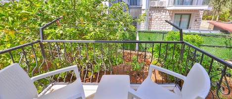 Un balcón con sillas y mesa de plástico blanco, rodeado de plantas y árboles de exuberante follaje.