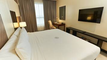 Quarto de hotel com uma cama grande, uma mesa com cadeira, televisão e janela com cortinas.