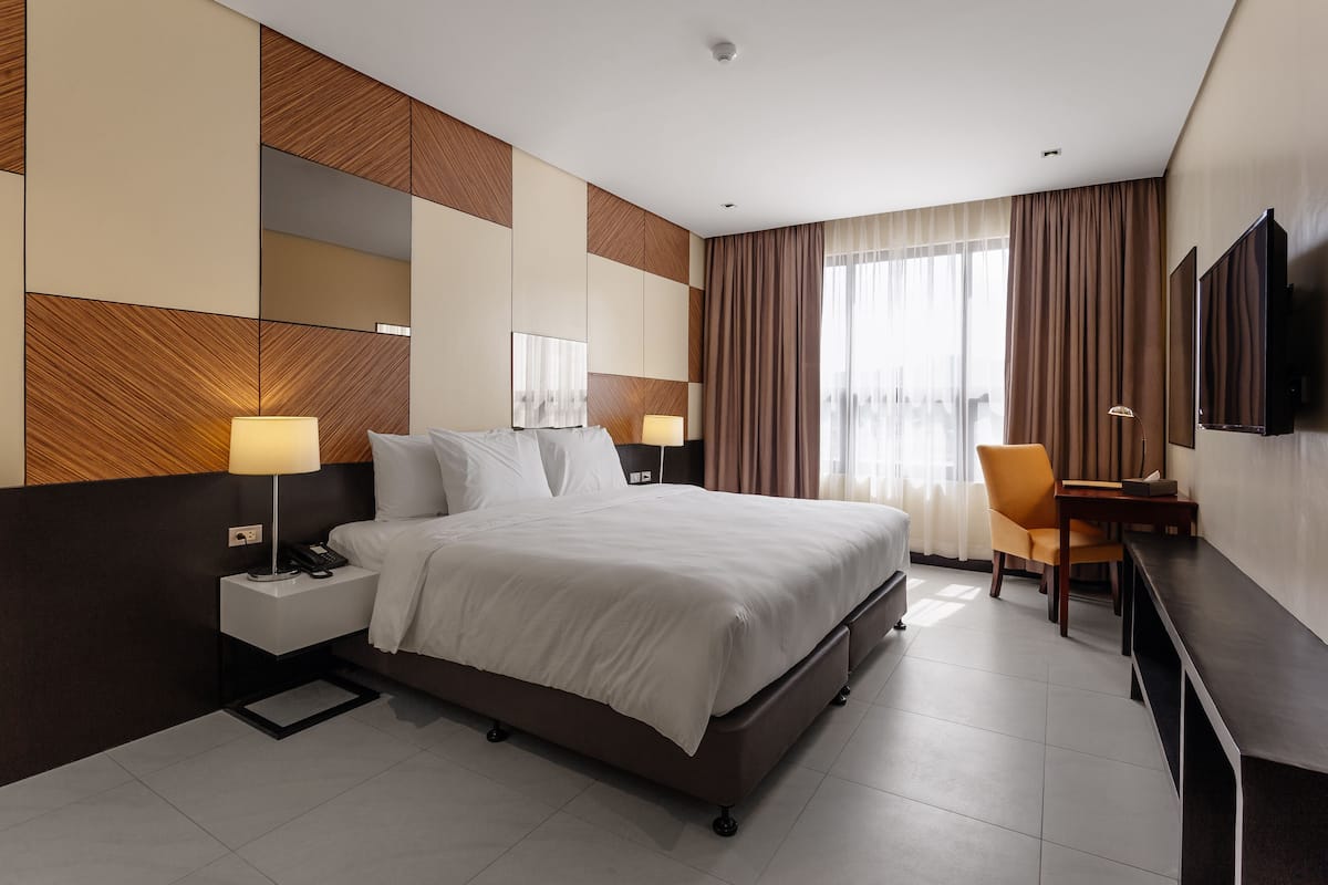 A modern hotel room with a large bed, bedside tables with lamps, a desk with a chair, and a flat-screen TV mounted on the wall.
