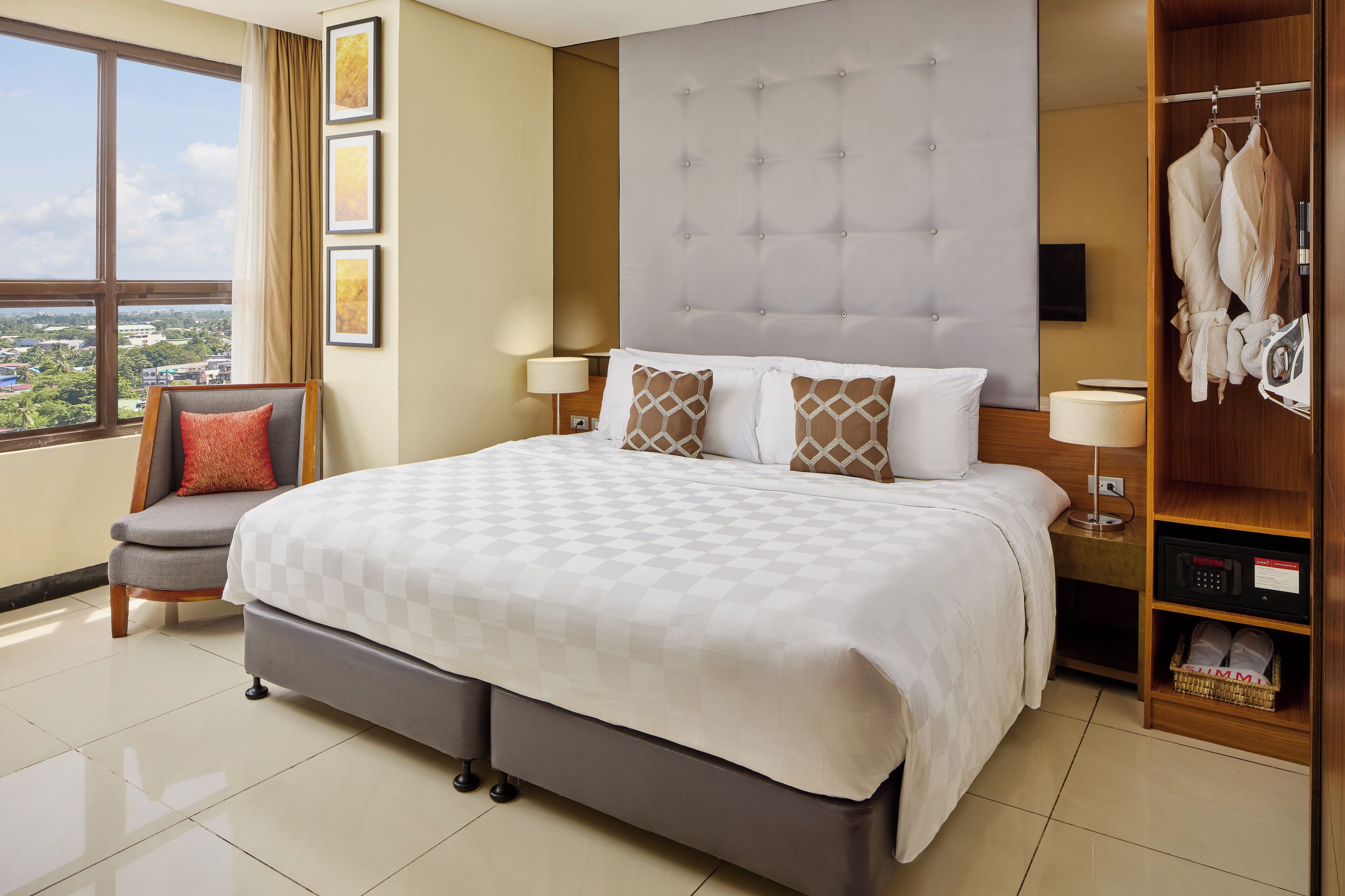 A hotel room with a large bed, a chair, a wardrobe, and a view of the city.