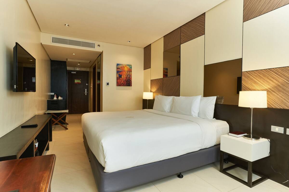 A hotel room with a large bed, bedside tables, a desk, a TV, and a painting on the wall.