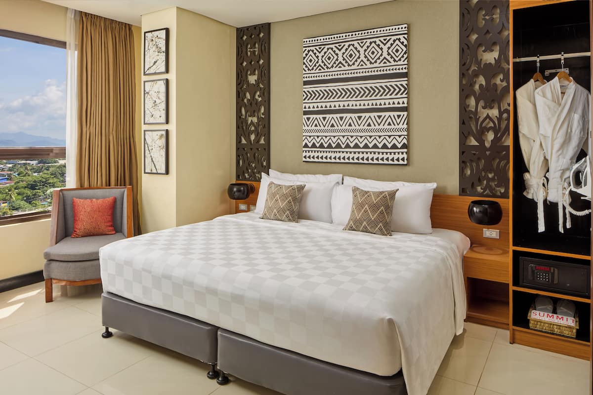 A hotel room with a large bed, a chair, a wardrobe, and a view of the city.