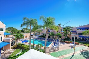 Un área de piscina con sillas de descanso y sombrillas, rodeada de palmeras y edificios.