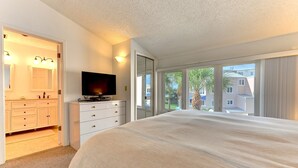 A hotel room with a large bed, a TV on a dresser, a bathroom with a mirror and sink, and a view of palm trees and buildings through the windows.