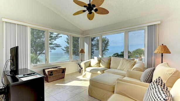 A spacious living room with a large window overlooking the sea, a ceiling fan, and a black entertainment unit.