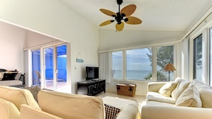 A spacious living room with a large window offering a view of the sea, a ceiling fan, and a television on a dark wooden stand.
