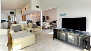 A modern living room with a flat-screen TV, a dining area with chairs, and a kitchen with a refrigerator.