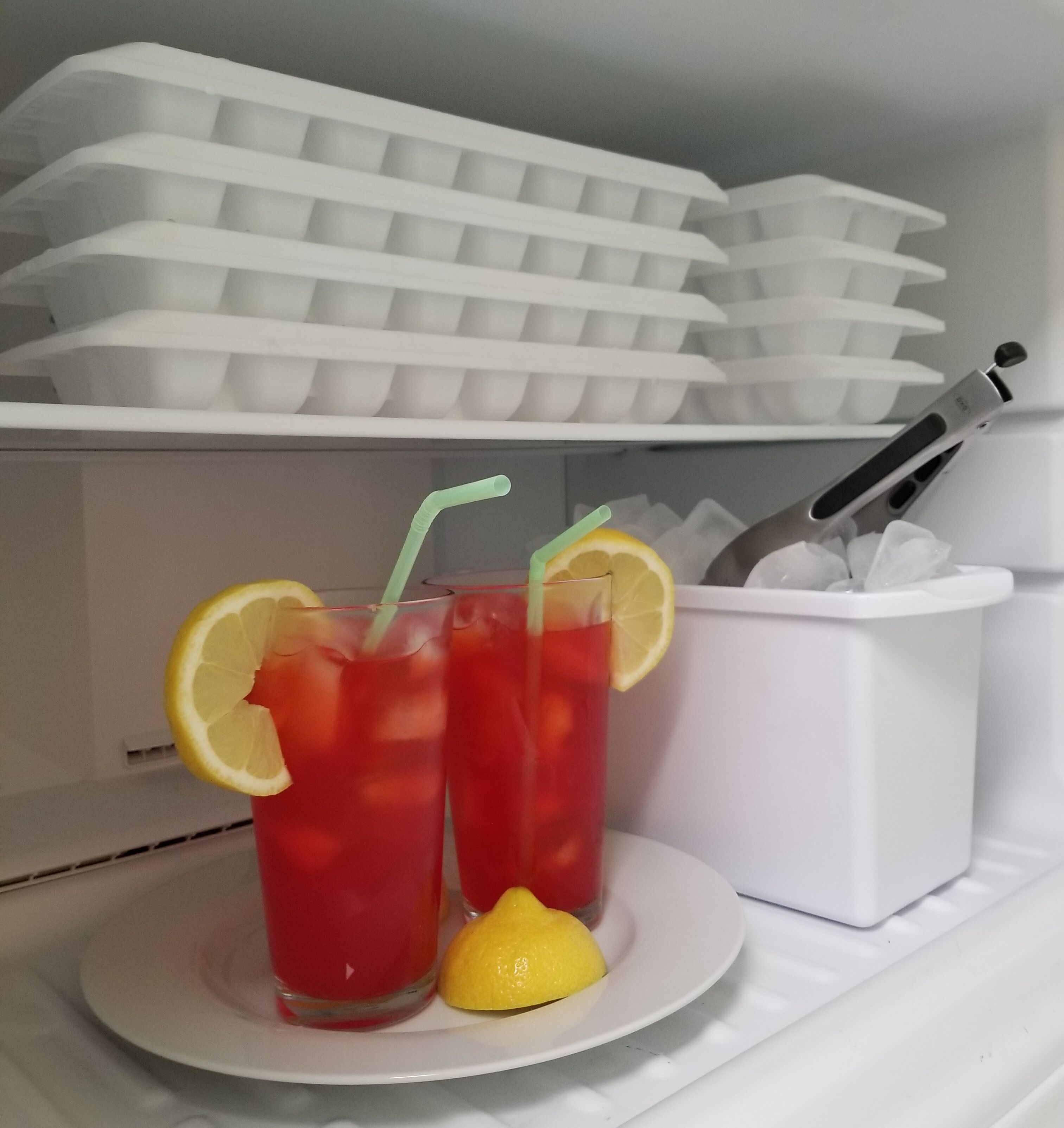 Dos vasos de bebida roja con rodajas de limón y pajitas sobre un plato blanco, un recipiente blanco con utensilios y un módulo de almacenamiento blanco con compartimentos.