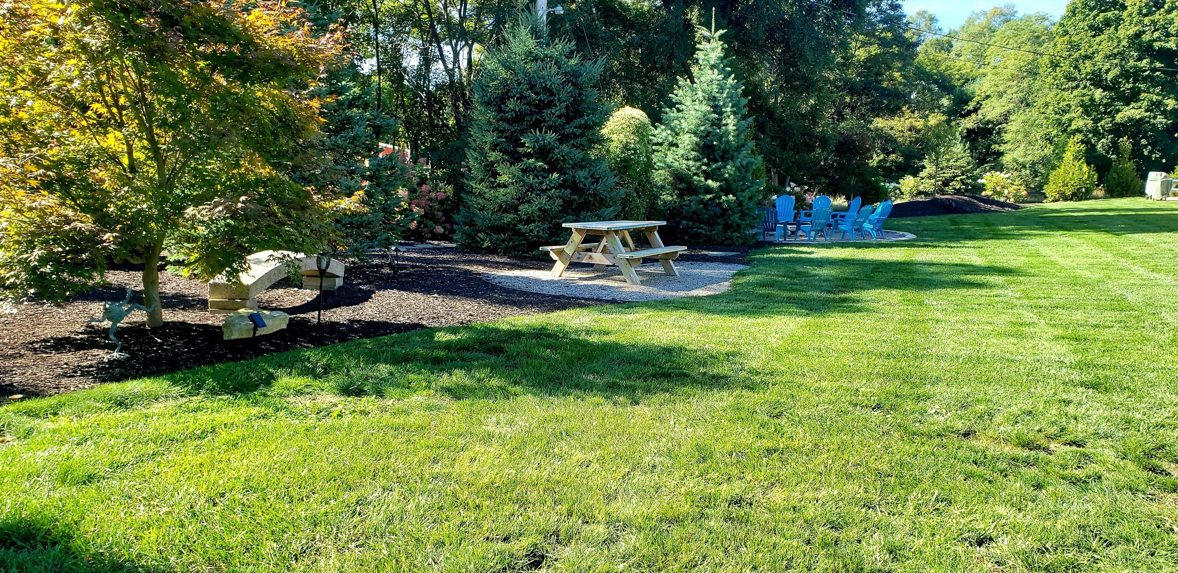 A picnic area with a wooden table and benches, surrounded by a grassy lawn and trees.