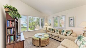 A living room with a sofa, a coffee table, a bookshelf, and a window with blinds. - Unit #305 Sand Cay Beach Resort Gulf View (Longboat Key)