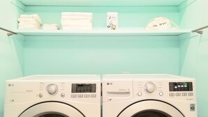 A laundry room with two white LG Sensor Dryers, a shelf with folded towels, a small bird figurine, and a hair dryer.