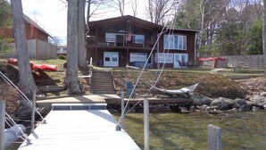 A cabin with a dock extending into a clear stream, surrounded by trees and a fenced yard with outdoor furniture. - Lake Winnipesaukee Waterfront! Family Friendly, Amazing View, HotTub,Kayaks+more (Alton Bay)