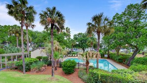 Une piscine entourée de palmiers et d’une végétation luxuriante, avec une terrasse en bois et des chaises longues.