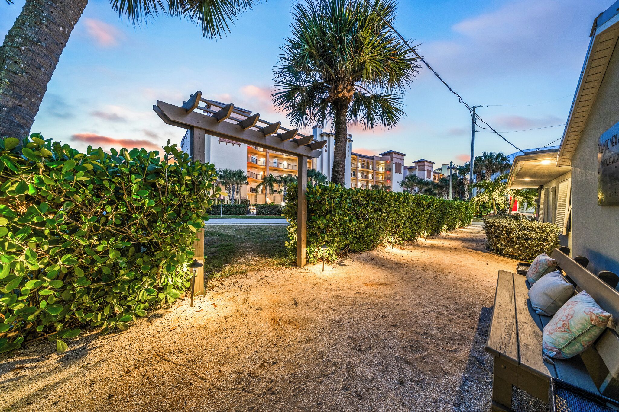 A pathway leading to a building with a pergola, surrounded by greenery and palm trees.