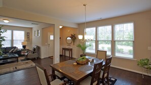 A dining area with a wooden table, chairs, and a chandelier. There is a sofa and a coffee table in the adjacent living room. - 3 Spacious Bedroom-Executive Town House, <br>Brand-New Construction, Duke RTP (Durham)