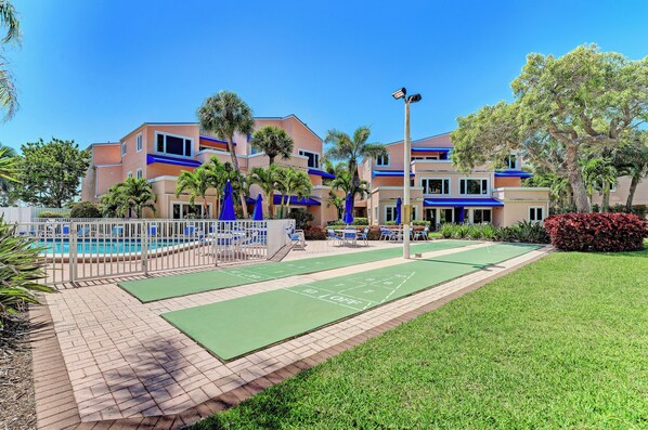 A pool area with a shuffleboard court, palm trees, and a building with blue accents. - Unit #302 Sand Cay Beach Resort Gulf Front (Longboat Key)