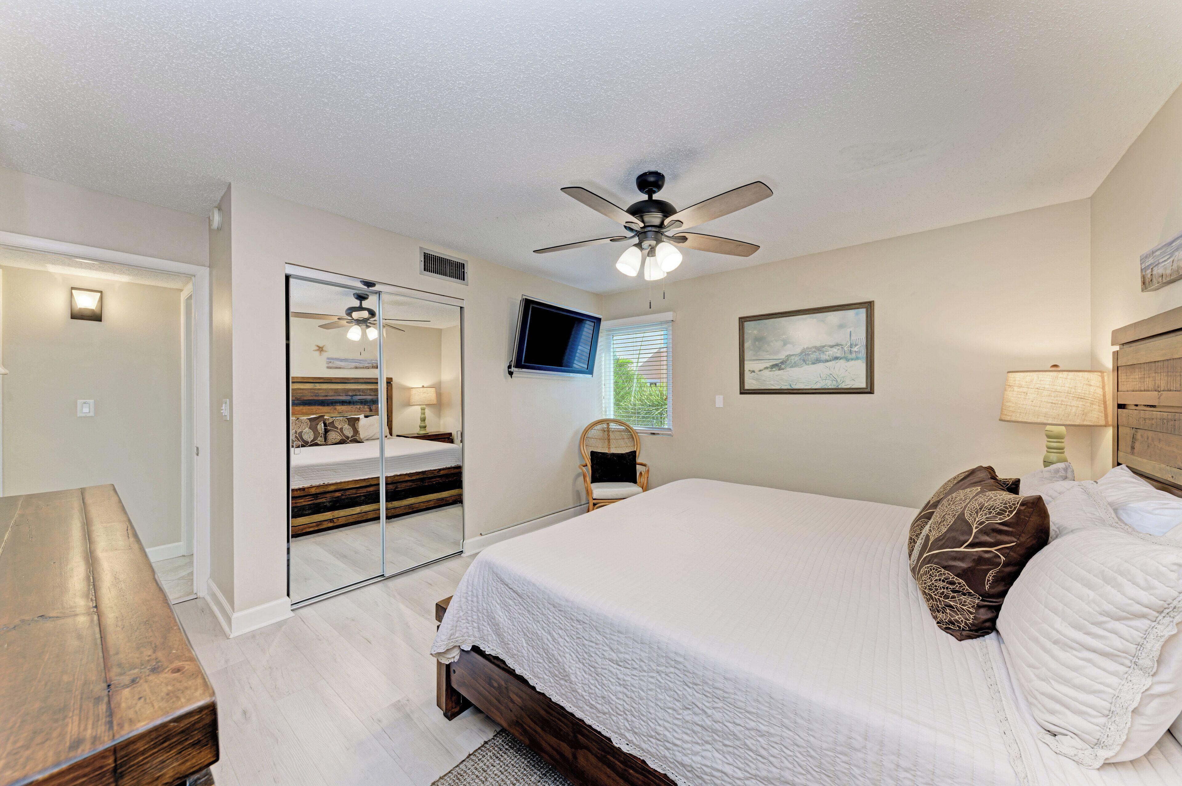 A bedroom with a large bed, a ceiling fan, a TV, a mirror, and a wooden bench.