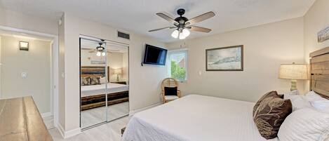 A bedroom with a large bed, a ceiling fan, a TV, a mirror, and a wooden bench.