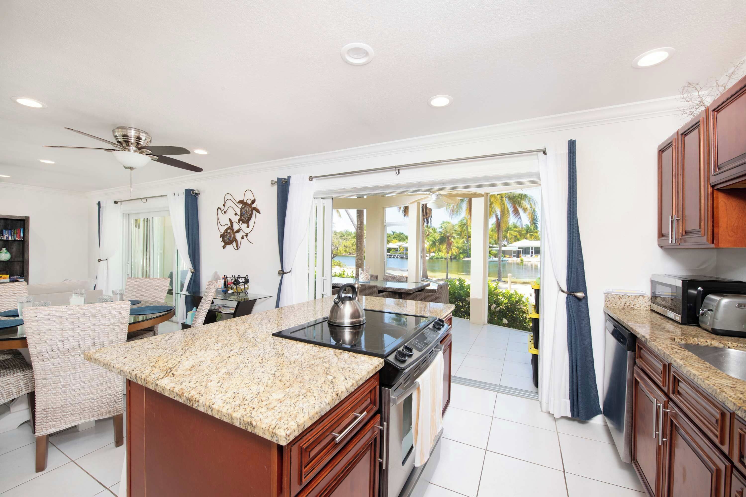 A kitchen with a central island, a dining area with wicker furniture, and a view of a pool and palm trees through the glass doors.