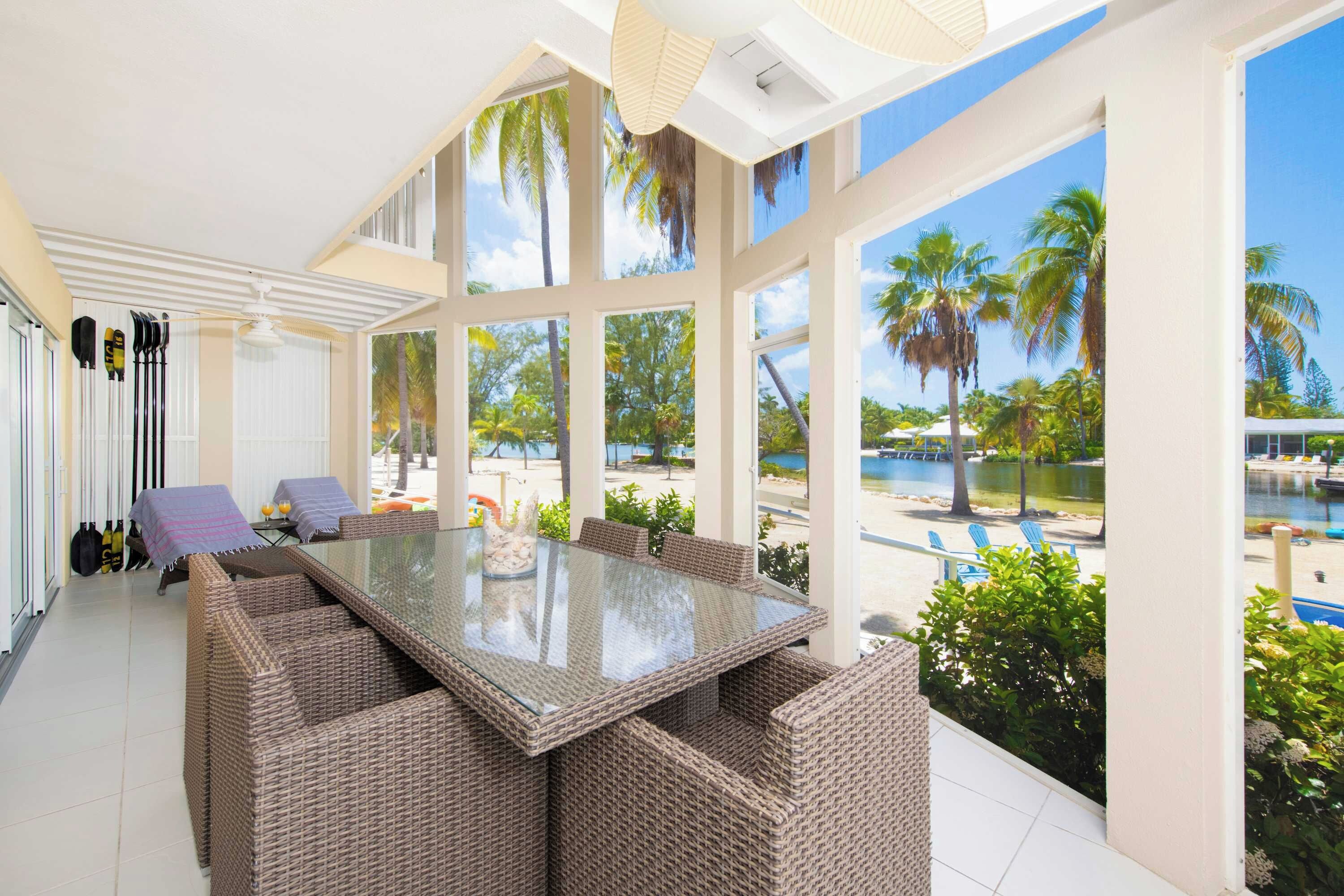 A sunlit room with a glass-top table surrounded by wicker chairs, large windows offering a view of palm trees and a pool.