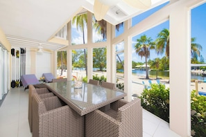 A sunlit room with a glass-top table surrounded by wicker chairs, large windows offering a view of palm trees and a pool.