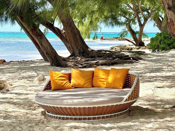 Una cama andadora de mimbre con cojines amarillos sobre una playa arenosa, rodeada de árboles y con vista al océano.