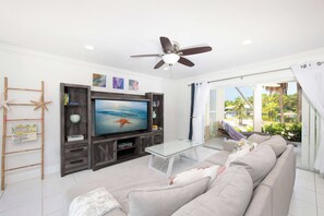 Ein Wohnzimmer mit einem Flachbildfernseher, einem Deckenventilator, einem Glastisch, einem beige farbenen Sofa und Blick auf einen Pool und Palmen durch das Fenster.