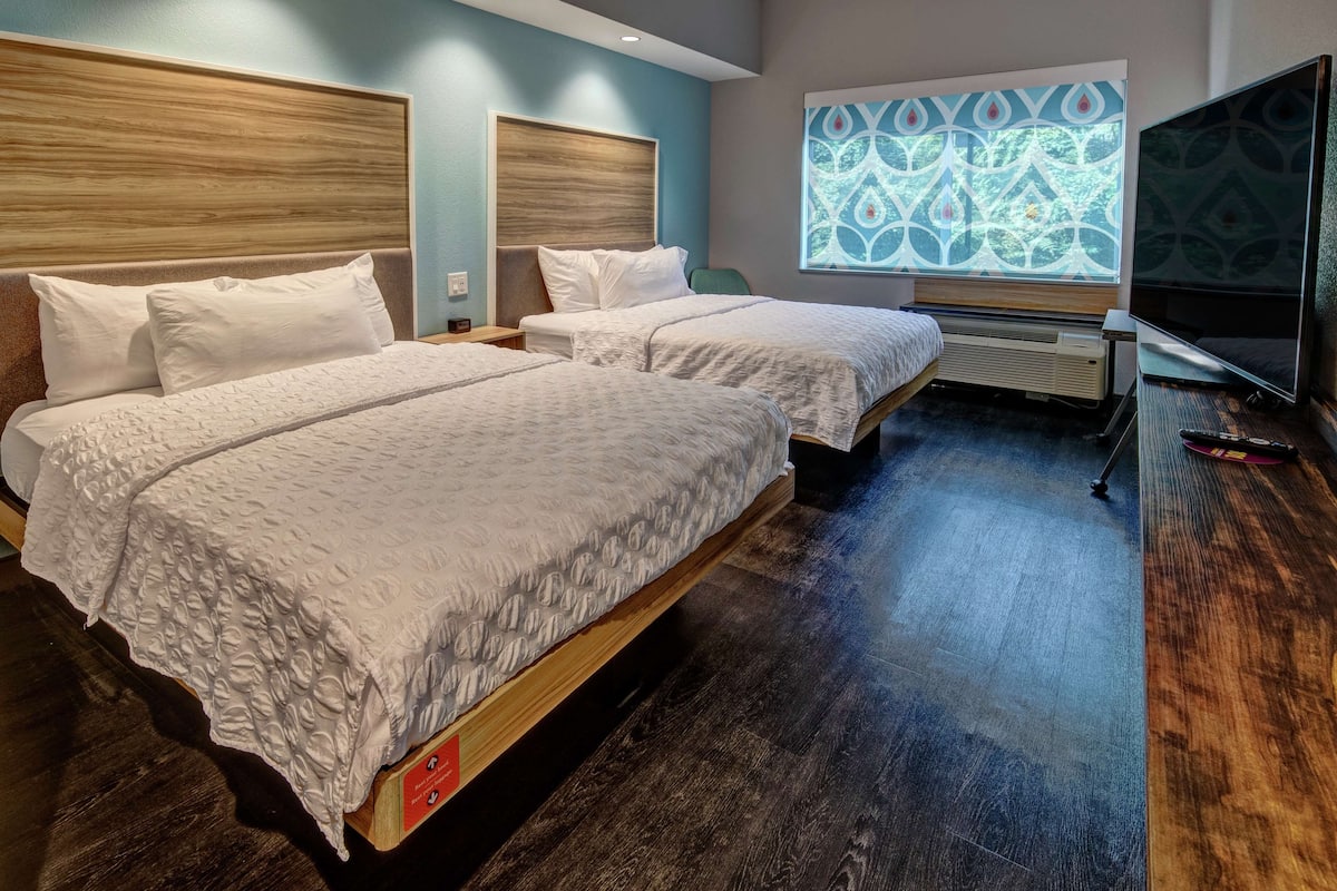 Ein Hotelzimmer mit zwei Betten, einem großen Fenster mit geometrischem Muster, einem Flachbildfernseher und Holzboden.