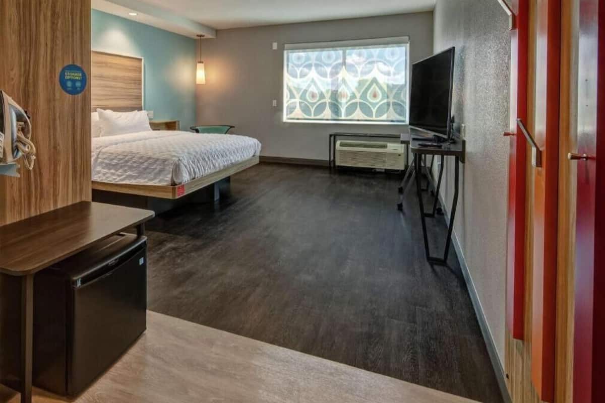 Ein Hotelzimmer mit einem Bett, einem Schreibtisch mit Computer, einem Fernseher und einem Fenster mit geometrischem Muster.