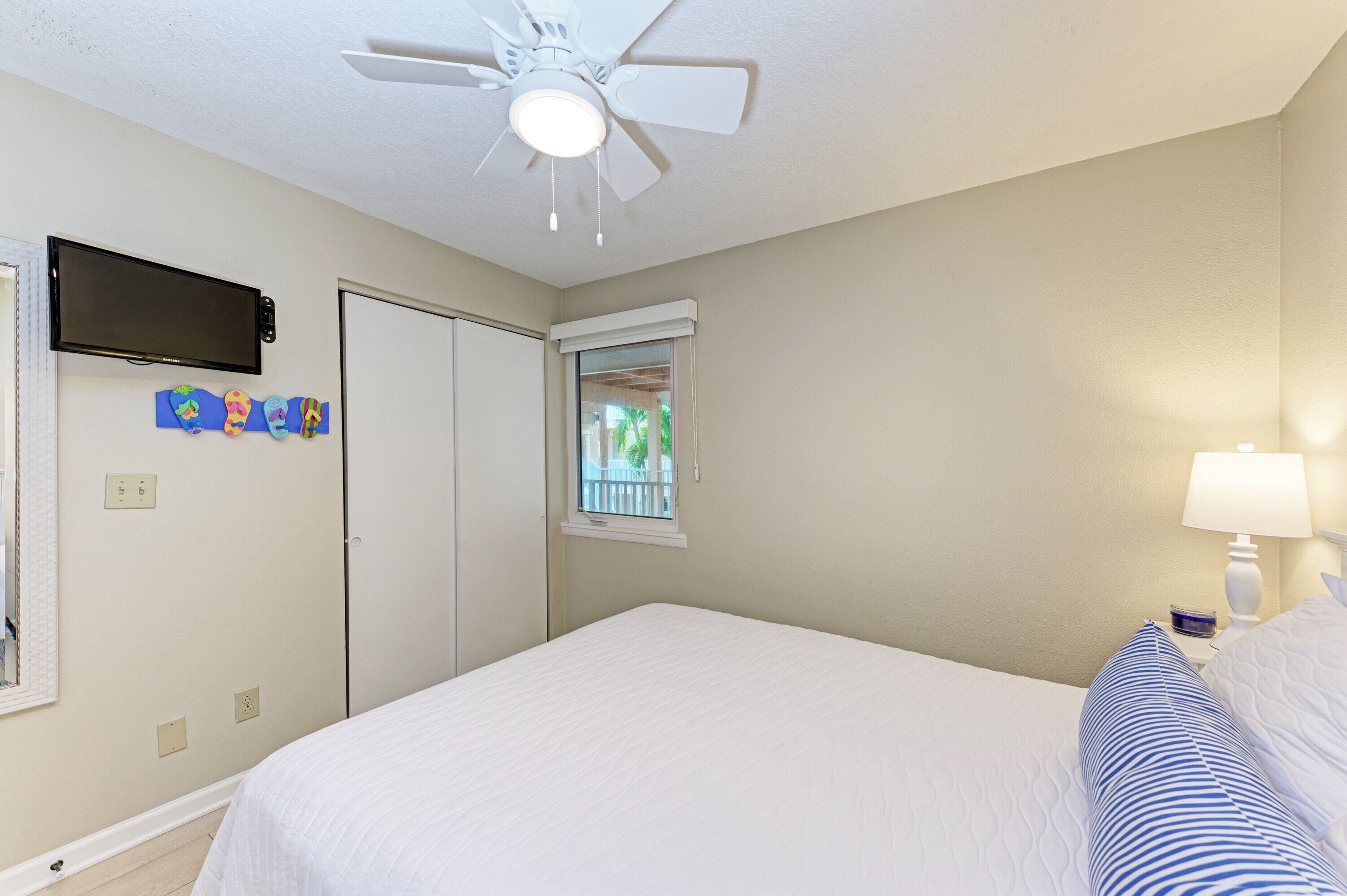 A bedroom with a bed, a ceiling fan, a TV, a window, and a nightstand with a lamp.