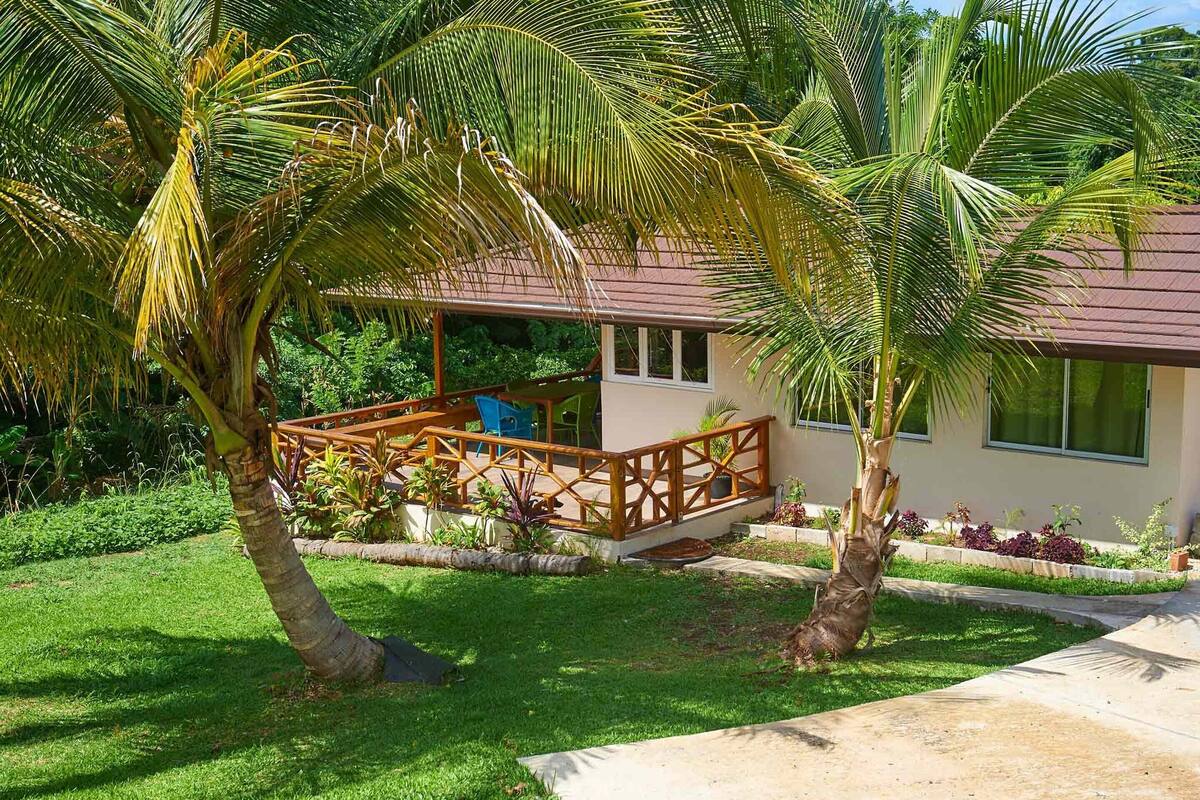 A house with a wooden deck, surrounded by palm trees and a well-maintained lawn.