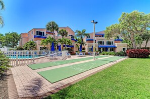 A pool area with a shuffleboard court, palm trees, and a building with blue accents.