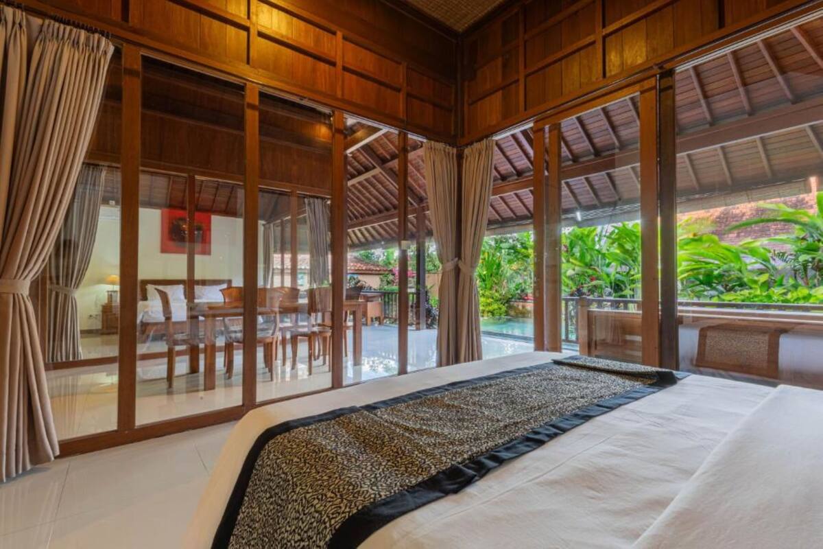 Un espacioso dormitorio con una cama grande, techo de madera y amplias ventanas que dan a un exuberante jardín.