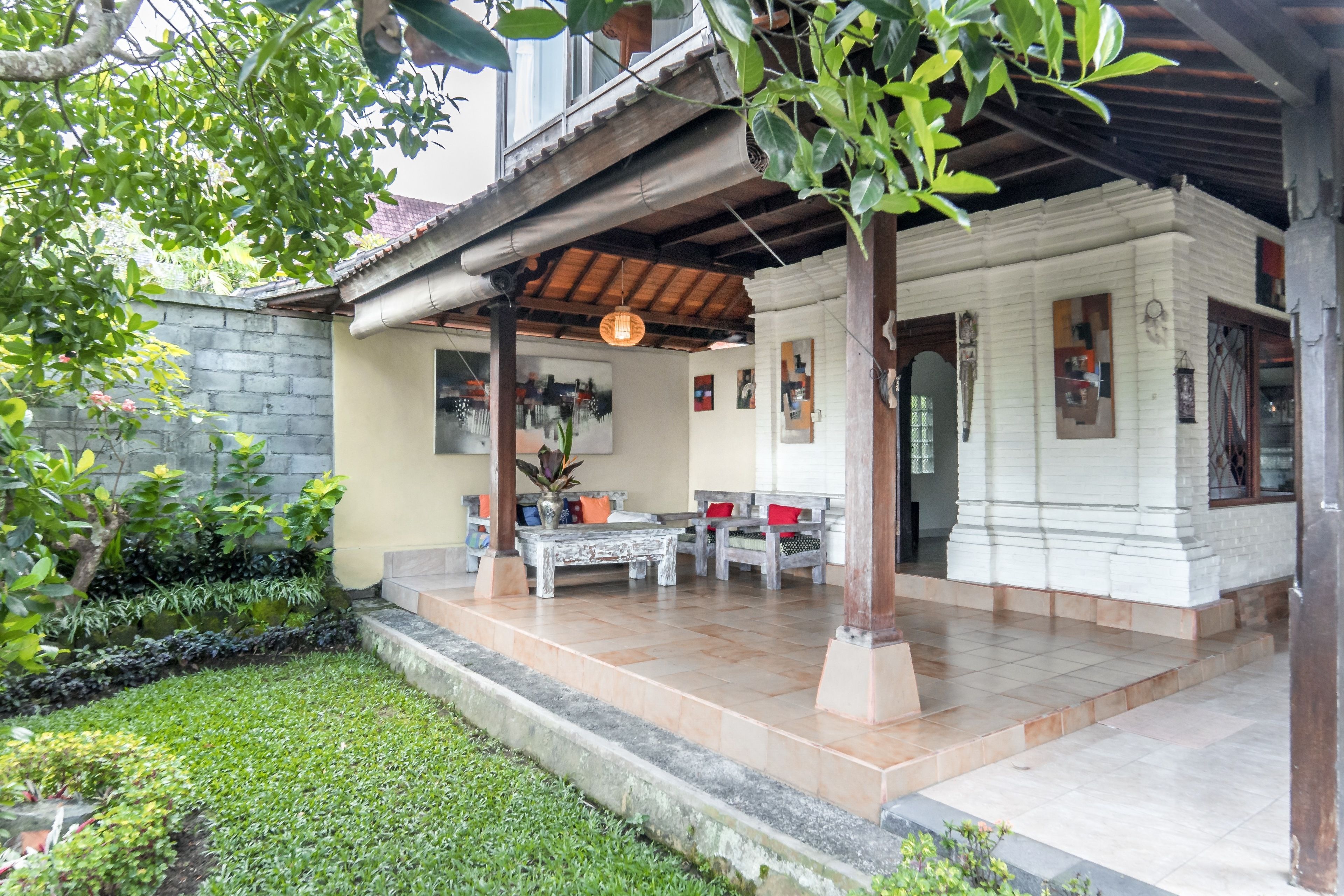 A covered outdoor seating area with a wooden pergola, a small white building with a tiled roof, and a garden with a stone wall and greenery.