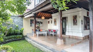 A covered outdoor seating area with a wooden pergola, a small white building with a tiled roof, and a garden with a stone wall and greenery.