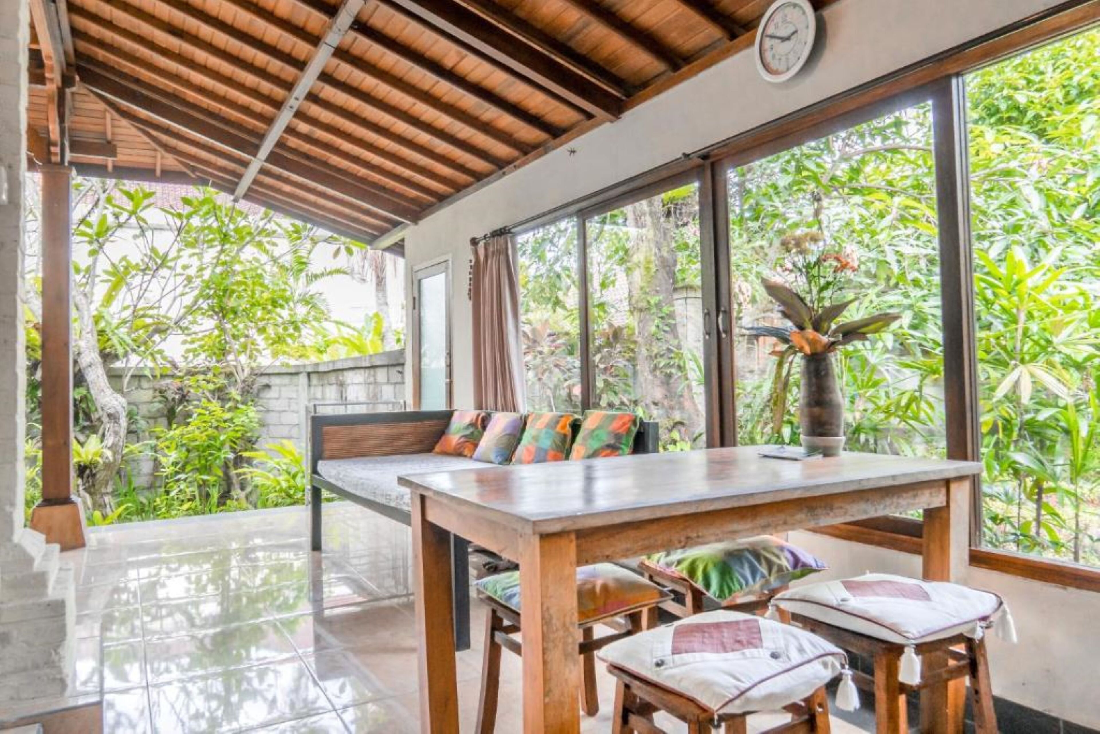 A dining area with a wooden table and chairs, a bench with colorful cushions, and large windows offering a view of lush greenery.