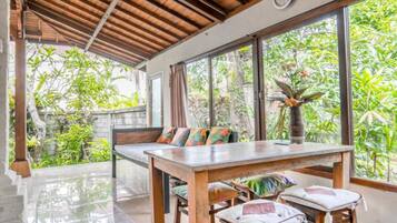 A dining area with a wooden table and chairs, a bench with colorful cushions, and large windows offering a view of lush greenery.