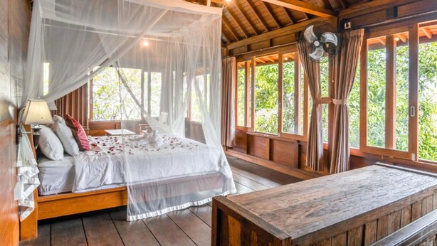 A wooden room with a bed under a canopy, large windows, and a wooden table. - Sari Bamboo Bungalows (Ubud)