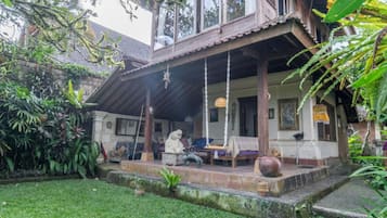 A traditional wooden house with a porch, surrounded by lush greenery.
