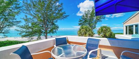 A terrace with a glass table and chairs overlooking a beach and ocean.